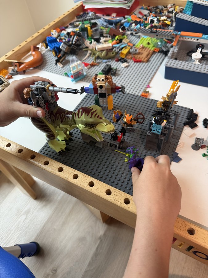 A child's hands mid-build at a LEGO table covered in sorted pieces, dinosaurs, and minifigures.
